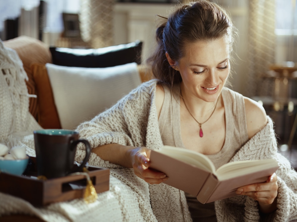 happy young woman in knitted cosy cardigan with tray and cup reading book at modern home in sunny autumn day.